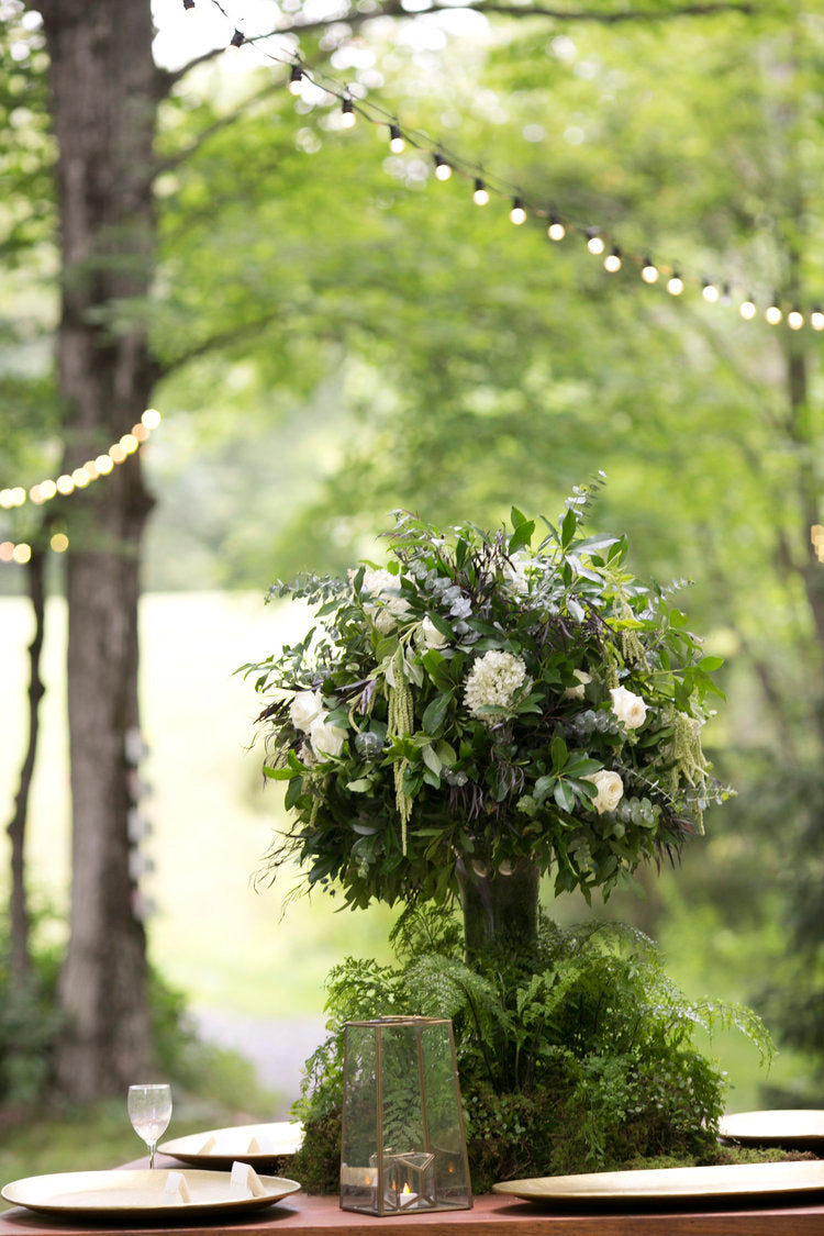 Decorative floral arrangement on a table outdoors with greenery and string lights.